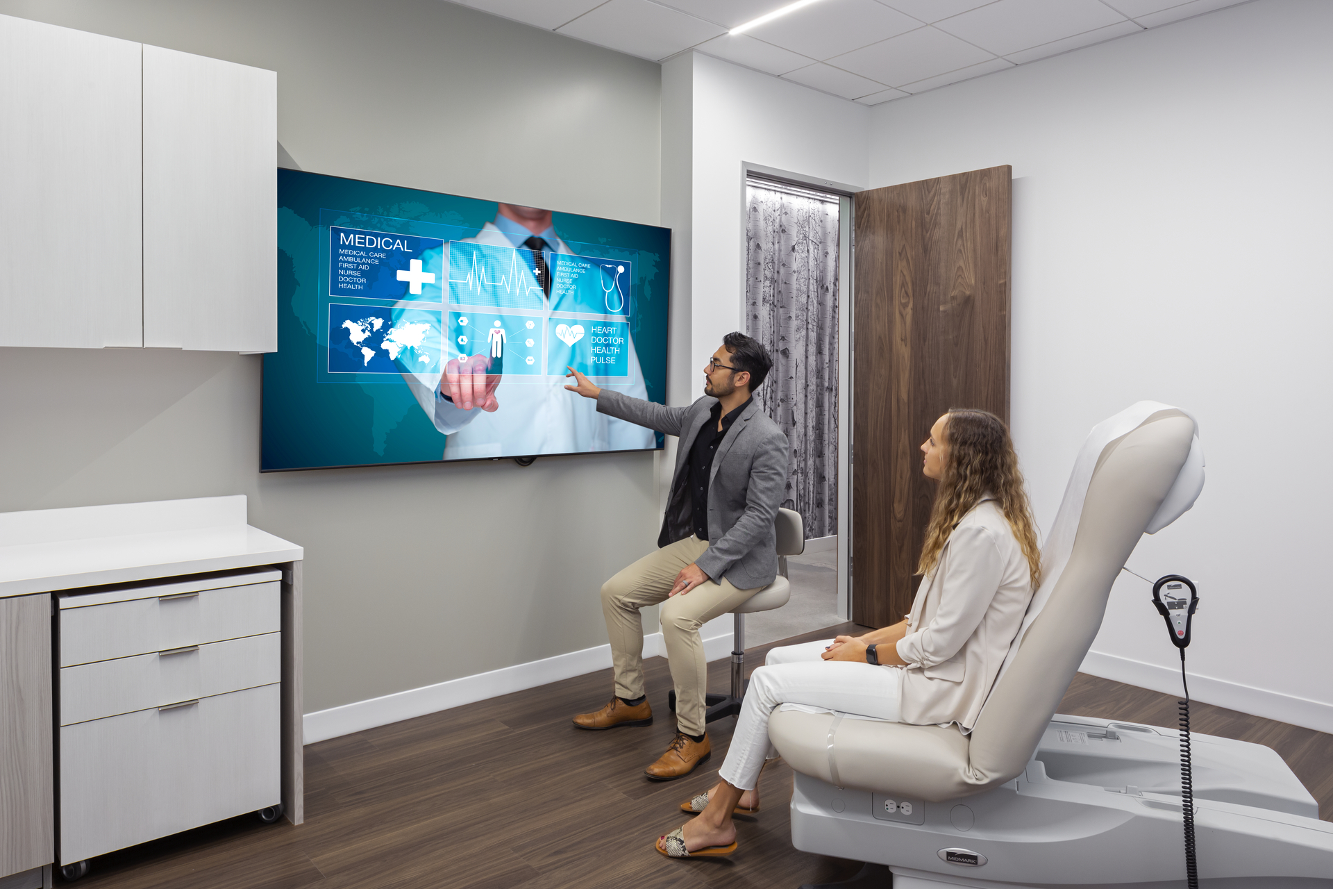This image shows a patient and a healthcare worker examining information on a large flat screen TV screen. They are in a patient's room and the patient and healthcare worker are both seated.